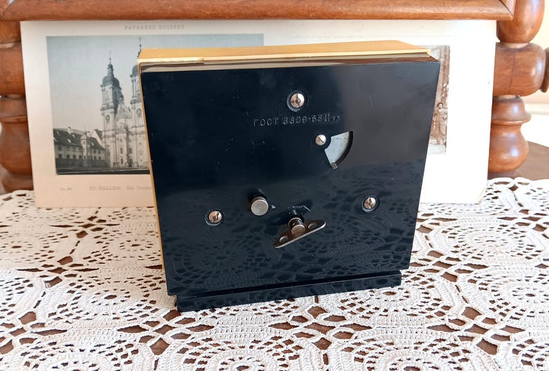 May include: A black music box with a gold trim. The music box has a small, round, silver knob on the front. The music box has a label on the front that reads 'root 8220-8311x'.