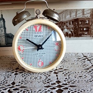 Può includere: Una sveglia bianca con un quadrante a motivi bianco e nero. L'orologio ha numeri rossi e le parole "Bevan" e "Made in Japan" sono stampate sul quadrante. L'orologio ha due campane argentate in cima.