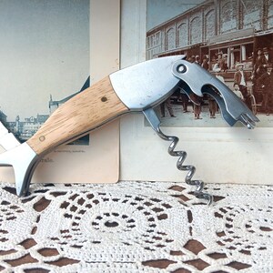 May include: A vintage fish-shaped corkscrew with a wooden handle and stainless steel components. The corkscrew features a blade and a bottle opener. The design is reminiscent of a fish, with the blade forming the tail.
