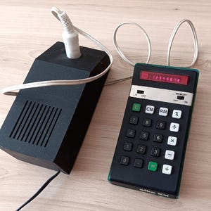 Peut inclure: Une calculatrice électronique noire avec des boutons blancs et verts, un écran numérique rouge affichant 12345678 et le texte elka 101. Elle est reliée par un cordon blanc à un bloc d'alimentation noir rectangulaire doté d'une grille de haut-parleur.