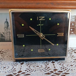 May include: A black and gold alarm clock with a black face and gold hands. The clock face has a black background with gold numbers and a gold second hand. The clock is sitting on a white crocheted surface. The clock face reads '12', '3', '6', and '9' and has the word 'Maas' printed on it.
