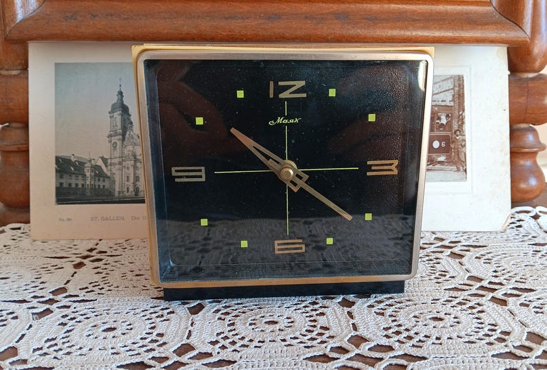 May include: A black and gold alarm clock with a black face and gold hands. The clock face has a black background with gold numbers and a gold second hand. The clock is sitting on a white crocheted surface. The clock face reads '12', '3', '6', and '9' and has the word 'Maas' printed on it.