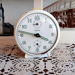 May include: White alarm clock with black hands and a green second hand. The clock face has black numbers and the word "GENFA" printed on it.
