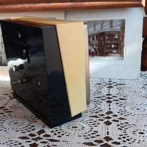 May include: A black and yellow alarm clock with a white face and silver hands. The clock is sitting on a white crocheted surface.