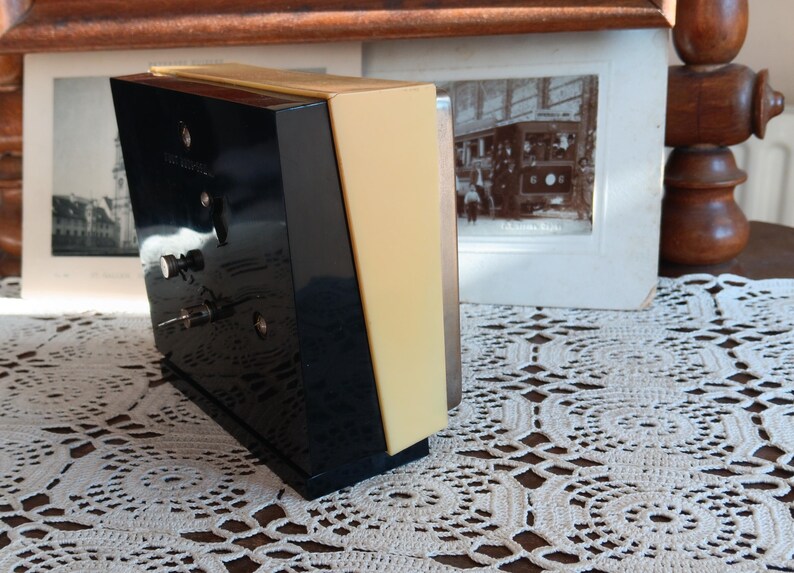 May include: A black and yellow alarm clock with a white face and silver hands. The clock is sitting on a white crocheted surface.