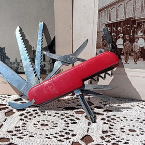 Può includere: Un coltello multiuso rosso con varie lame e strumenti in acciaio inossidabile, tra cui un coltello, forbici e una sega. Lo strumento è aperto, mostrando le sue funzioni. Lo sfondo include fotografie d'epoca e una tovaglia di pizzo bianco.