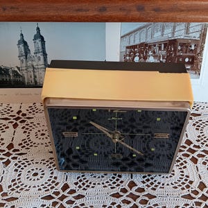 May include: A black and gold alarm clock with a black face and white hands. The clock is on a white crocheted surface. The clock face has the word 'Maxima' printed on it.