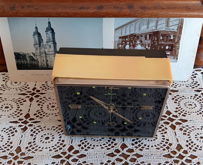May include: A black and gold alarm clock with a black face and white hands. The clock is on a white crocheted surface. The clock face has the word 'Maxima' printed on it.