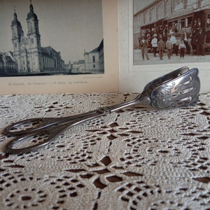 Puede incluir: Pinzas de servir plateadas con un diseño ornamentado. Las pinzas están abiertas y descansan sobre un mantel de ganchillo blanco.