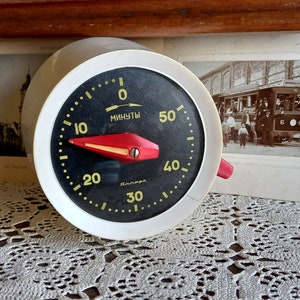 May include: A white kitchen timer with a black face and red needle. The timer has a black and yellow dial with numbers from 0 to 60. The dial is labelled "МИНУТЫ" (Russian for "minutes").