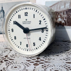Puede incluir: Un reloj analógico blanco con agujas negras y números negros. La esfera del reloj tiene el texto "JUNGHANS electronic" y "ATO-MAT" impreso en él.