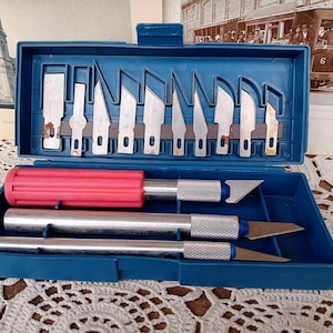 May include: A blue plastic craft knife set with a variety of blades and handles. The set includes a red handle and two silver handles with blades. The blades are stored in the lid of the case.