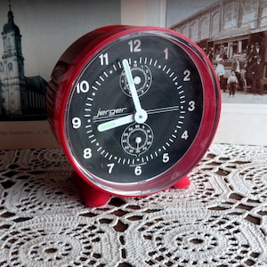 May include: A red alarm clock with a black face and white numbers. The clock has a second hand and a small dial for setting the alarm. The clock is made by Jerger.