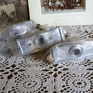 May include: Three silver-colored metal objects wrapped in clear plastic packaging. The objects are rectangular in shape and have a circular opening in the center. The objects are sitting on a white crocheted tablecloth.