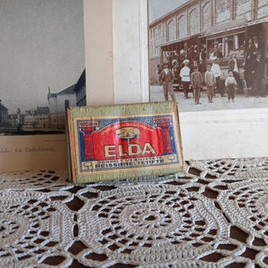 Puede incluir: Una caja de cerillas vintage con un diseño verde y rojo. La caja de cerillas está etiquetada con "ELDA" y dice "100 Stück" y "UNDURCHDRÜCKBAR" en la parte delantera. La caja de cerillas está sobre un encaje blanco de ganchillo.