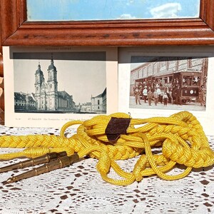 May include: A vintage display featuring a yellow braided rope with brass-colored metal tips, a brown leather accent, and two antique black and white photographs. The photographs depict a church and a street scene.