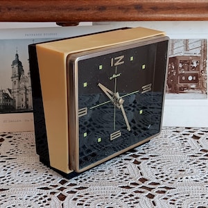 May include: A square, cream-colored alarm clock with a black face. The clock has gold-colored hands and a gold-colored bezel. The clock face has black numbers and the word 'IZ' in the top center. The clock is sitting on a white crocheted surface.