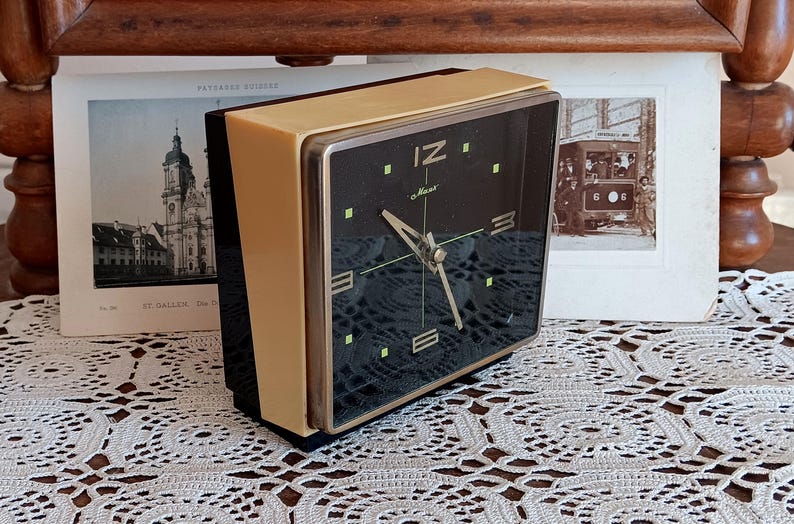 May include: A square, cream-colored alarm clock with a black face. The clock has gold-colored hands and a gold-colored bezel. The clock face has black numbers and the word 'IZ' in the top center. The clock is sitting on a white crocheted surface.