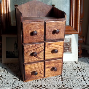 Puede incluir: Un pequeño armario de madera con seis cajones, cada uno con una perilla marrón. El armario está teñido de un color marrón oscuro y tiene un aspecto ligeramente desgastado. El armario está sentado sobre un mantel de encaje blanco.