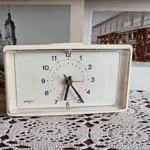 May include: White analog clock with black hands and a white face. The clock has black numbers and a black 'wigo' logo. The clock is sitting on a white lace tablecloth.