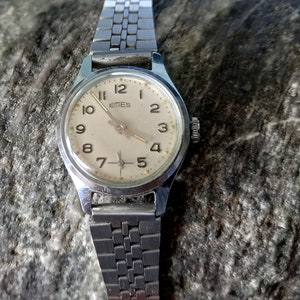 Peut inclure: Une montre-bracelet argentée avec un cadran blanc et des chiffres noirs. La montre a un bracelet en métal argenté et indique "Emes" sur le cadran.