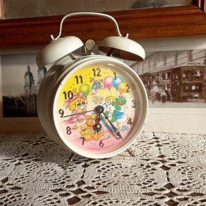 May include: White alarm clock with a colorful face featuring characters from the Muppet Babies. The clock has a pink background with a green frog, a pink pig, and a yellow duck. The clock face has black numbers and hands.