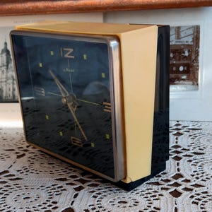 May include: A black and yellow alarm clock with a black face and white numbers. The clock has a silver rim around the face and a black plastic back. The clock is sitting on a white lace tablecloth.