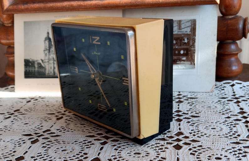 May include: A black and yellow alarm clock with a black face and white numbers. The clock has a silver rim around the face and a black plastic back. The clock is sitting on a white lace tablecloth.