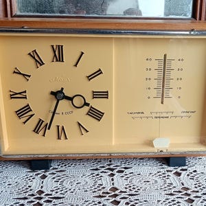 Puede incluir: Un reloj vintage enmarcado en madera con una esfera blanca y marcadores de hora en números romanos negros. El reloj también tiene un termómetro con una aguja negra y un fondo blanco. La esfera del reloj dice "Сделано в СССР" que se traduce como "Hecho en la URSS".