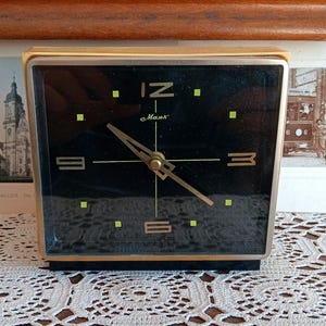 May include: A black and gold alarm clock with a black face and gold hands. The clock has a gold frame and is set to 12:00. The clock face has the words 'C. Maas' printed on it. The clock is sitting on a white lace tablecloth.