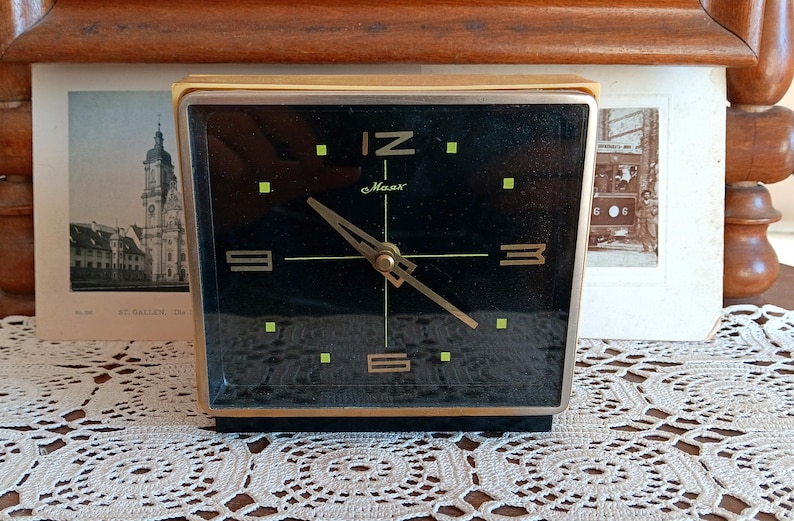 May include: A black and gold alarm clock with a black face and gold hands. The clock has a gold frame and is set to 12:00. The clock face has the words 'C. Maas' printed on it. The clock is sitting on a white lace tablecloth.