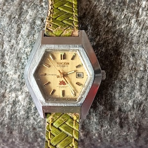 Op de afbeelding: Een zilverkleurig polshorloge met een gouden wijzerplaat en zwarte wijzers. Het horloge heeft een groene geweven band en staat "Ricoh Automatic Crystal" op de wijzerplaat.