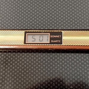 Puede incluir: Un reloj digital dorado con una esfera negra y las palabras "Sonnet Quartz" impresas en él. El reloj muestra la hora "5:01".