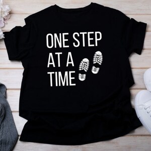 May include: Black t-shirt with white text that reads "One Step At A Time" and two white footprints.
