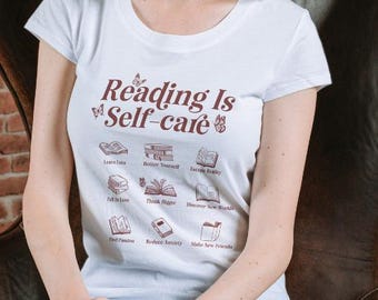 May include: White t-shirt with the text "Reading Is Self-care" in brown lettering. The shirt features illustrations of books with text such as "Learn Local", "Better Yourself", "Escape Reality", "Fall in Love", "Think Bigger", "Discover New Worlds", "Find Passion", "Reduce Anxiety", and "Make New Friends".