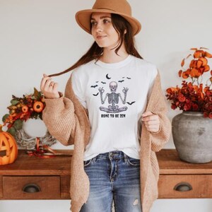 Peut inclure: T-shirt blanc avec un motif de squelette en position de yoga, le texte dit "BONE TO BE ZEN". Le motif comprend un croissant de lune et des chauves-souris. La personne porte un chapeau marron et un cardigan beige.
