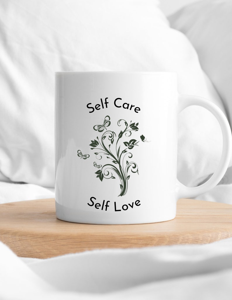 May include: White ceramic mug with a black and green design featuring a stylized tree with butterflies and the text "Self Care Self Love".