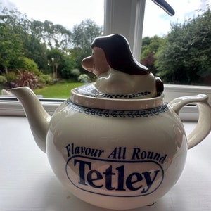 May include: A white ceramic teapot with a blue and white Tetley logo that says "Flavour All Round Tetley". The teapot has a brown and white dog figurine on the lid.