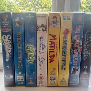 Puede incluir: Una colección de cintas VHS con títulos de películas como "Santa Claus", "Stuart Little 2", "The Grinch", "Matilda" y "Casper". Las cintas están dispuestas verticalmente, con etiquetas coloridas e ilustraciones de películas.