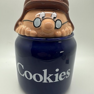 Op de afbeelding: Een blauwe keramische koekjespot met een wit "Cookies"-label. De deksel is gevormd als een cartoonman met bruin haar, een bril en een bruine hoed.