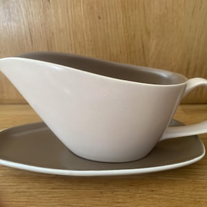 Può includere: Una salsiera con un esterno liscio color crema chiaro e un interno color tortora a contrasto. Il recipiente presenta un manico curvo e un beccuccio appuntito, poggiato su un piattino ovale abbinato color tortora con bordo color crema.