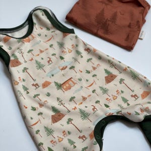 Könnte beinhalten: Ein cremefarbener Baby-Strampler mit einem Camping-Motiv mit Zelten, Bäumen und Hirschen. Der Strampler hat dunkelgrüne Paspeln und Druckknöpfe. Ein rostfarbener Body mit einer Waldszene ist ebenfalls vorhanden.
