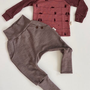 Könnte beinhalten: Ein gestreiftes Langarmshirt mit rosa Grund und schwarzen Streifen. Das Shirt hat einen Katzendruck und den Text "Little Wild One" unten rechts. Eine gestreifte Hose mit braunem Grund und weißen Streifen.