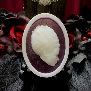 May include: A white cameo with a profile of a person's head, set in a purple oval frame with a white border. The cameo is on a black stand.
