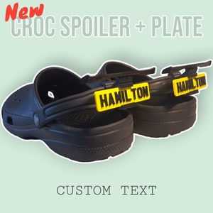 Puede incluir: Un par de Crocs negras con placas de nombre "HAMILTON" amarillas y negras unidas a las correas traseras. Las placas de nombre están unidas con un clip de plástico negro. Las Crocs están sobre un fondo verde claro. Se puede personalizar el texto.