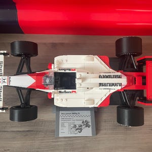 K&ouml;nnte beinhalten: Ein wei&szlig;es und rotes Lego-Modell eines McLaren MP4/4 Formel-1-Rennwagens mit Marlboro- und Shell-Branding. Das Auto hat schwarze R&auml;der und ein schwarzes Chassis. Das Auto steht auf einer wei&szlig;en Oberfl&auml;che.