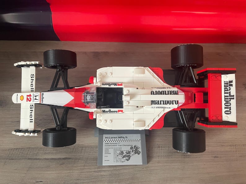 K&ouml;nnte beinhalten: Ein wei&szlig;es und rotes Lego-Modell eines McLaren MP4/4 Formel-1-Rennwagens mit Marlboro- und Shell-Branding. Das Auto hat schwarze R&auml;der und ein schwarzes Chassis. Das Auto steht auf einer wei&szlig;en Oberfl&auml;che.