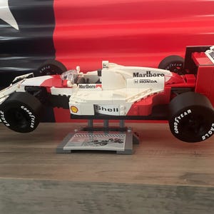 K&ouml;nnte beinhalten: Ein rotes, wei&szlig;es und schwarzes Lego-Modell eines Formel-1-Rennwagens mit den Worten "Marlboro" und "Shell" an der Seite. Das Auto steht auf einem grauen St&auml;nder mit einem schwarz-wei&szlig;en Schachbrettmuster.