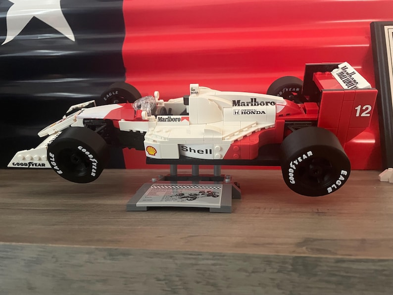 K&ouml;nnte beinhalten: Ein rotes, wei&szlig;es und schwarzes Lego-Modell eines Formel-1-Rennwagens mit den Worten "Marlboro" und "Shell" an der Seite. Das Auto steht auf einem grauen St&auml;nder mit einem schwarz-wei&szlig;en Schachbrettmuster.