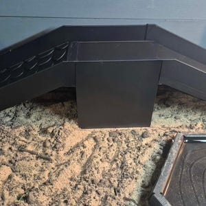 May include: A matte black metal sluice box featuring a central rectangular collection chamber with two angled side channels. The left channel includes a series of raised riffles for material trapping, while the right channel is smooth.